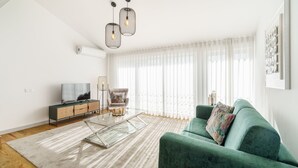 A modern living room with a green sofa, a glass coffee table, a TV on a wooden cabinet, and a large window with sheer curtains. - Souto Essence Apartment By Minhos Guest (Braga)