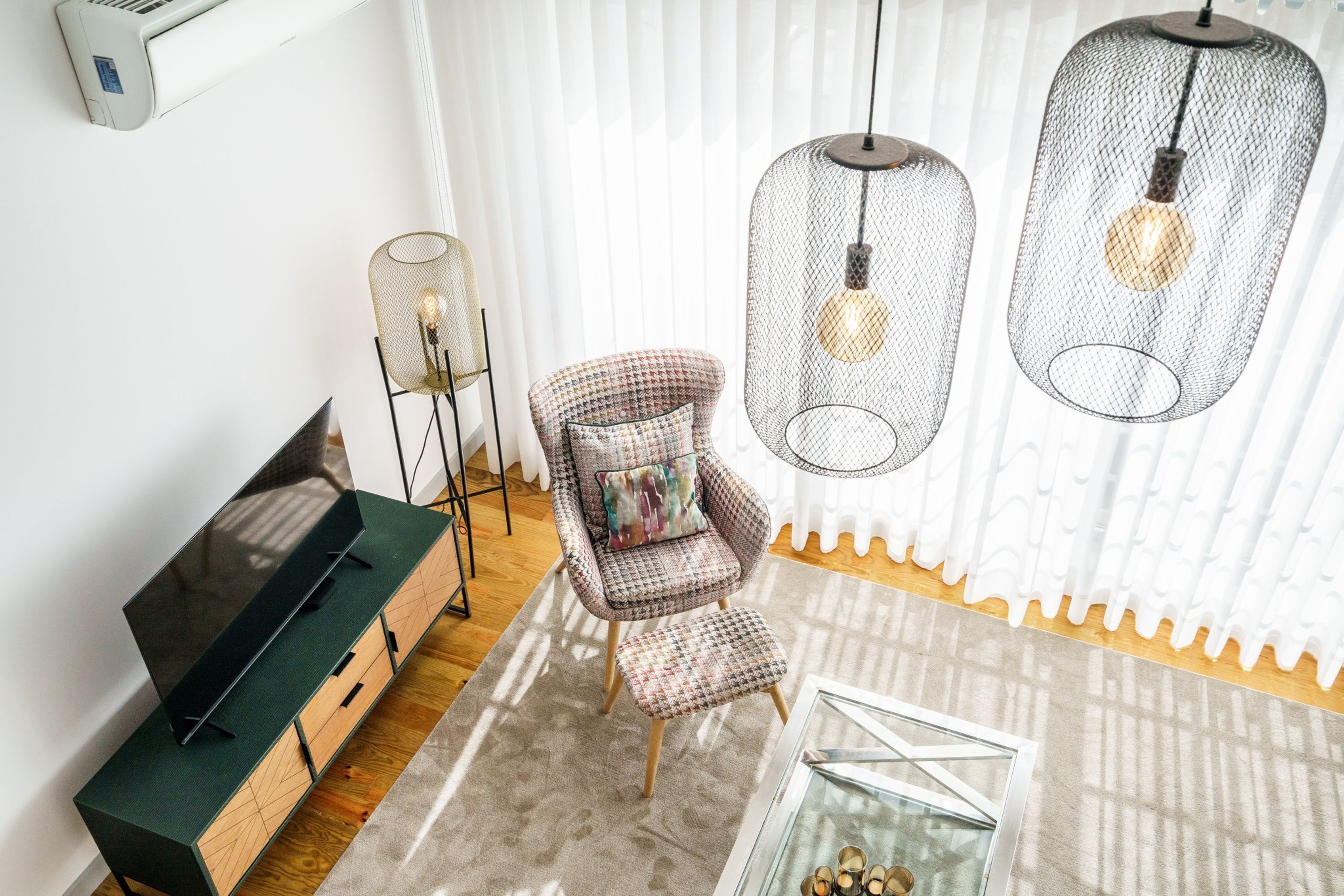 A modern living room with a flat-screen TV on a wooden cabinet, a plaid armchair with a cushion, and a glass coffee table with shoes.