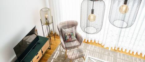 A modern living room with a flat-screen TV on a wooden cabinet, a plaid armchair with a cushion, and a glass coffee table with shoes.