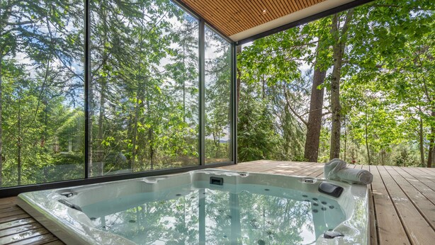 Ein Whirlpool mit einer Holzterrasse und großen Fenstern mit Blick auf einen Wald.