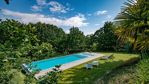 Ein rechteckiger Swimmingpool mit zwei Liegestühlen und einem kleinen Tisch auf einer grünen Rasenfläche, umgeben von dichtem Grün.