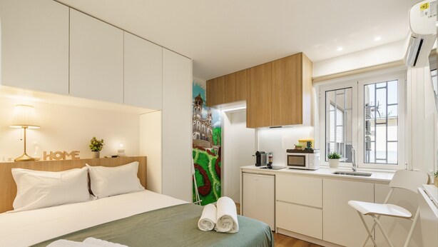 A compact hotel room with a bed, a small kitchenette, a window, and a wall-mounted air conditioner. - Liberty Braga Apartment by Minho s Guest (Braga)