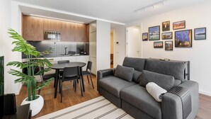 A modern living room with a grey sofa, a dining table with chairs, a kitchen area with wooden cabinets, and a potted plant. - 196 Andrade Corvo Apartment by Minho s Guest (Braga)