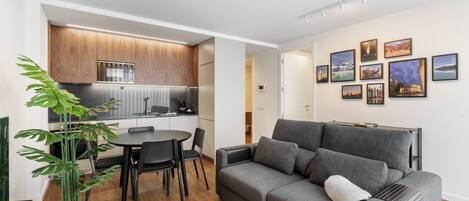 A modern living room with a grey sofa, a dining table with chairs, a kitchen area with wooden cabinets, and a potted plant.