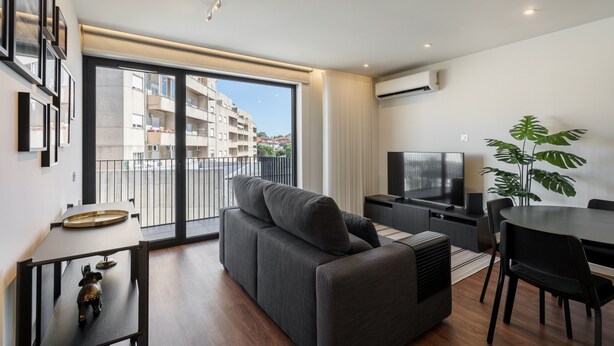 A modern living room with a sofa, TV, dining table, and large windows overlooking a balcony. - 196 Andrade Corvo Apartment by Minho s Guest (Braga)