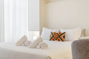 Un lit bien fait, avec des serviettes pliées, un coussin coloré et une tête de lit blanche, dans une chambre d’hôtel.