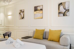 Une chambre d’hôtel moderne avec un canapé gris, deux coussins de couleur moutarde, une table basse en verre et deux serviettes enroulées.