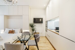Ein modernes Hotelzimmer mit Bett, Glastisch, zwei Stühlen, einer kleinen Topfpflanze, einem an der Wand montierten Fernseher und einer Küchenzeile mit Spüle und Arbeitsplatte.