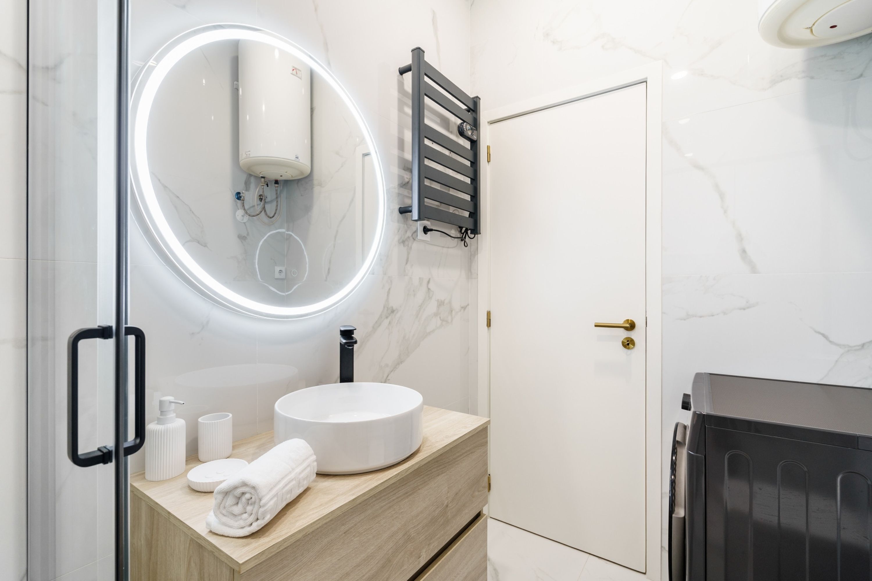 A modern bathroom with a round mirror, a towel dispenser, a toilet paper holder, and a towel on the counter.
