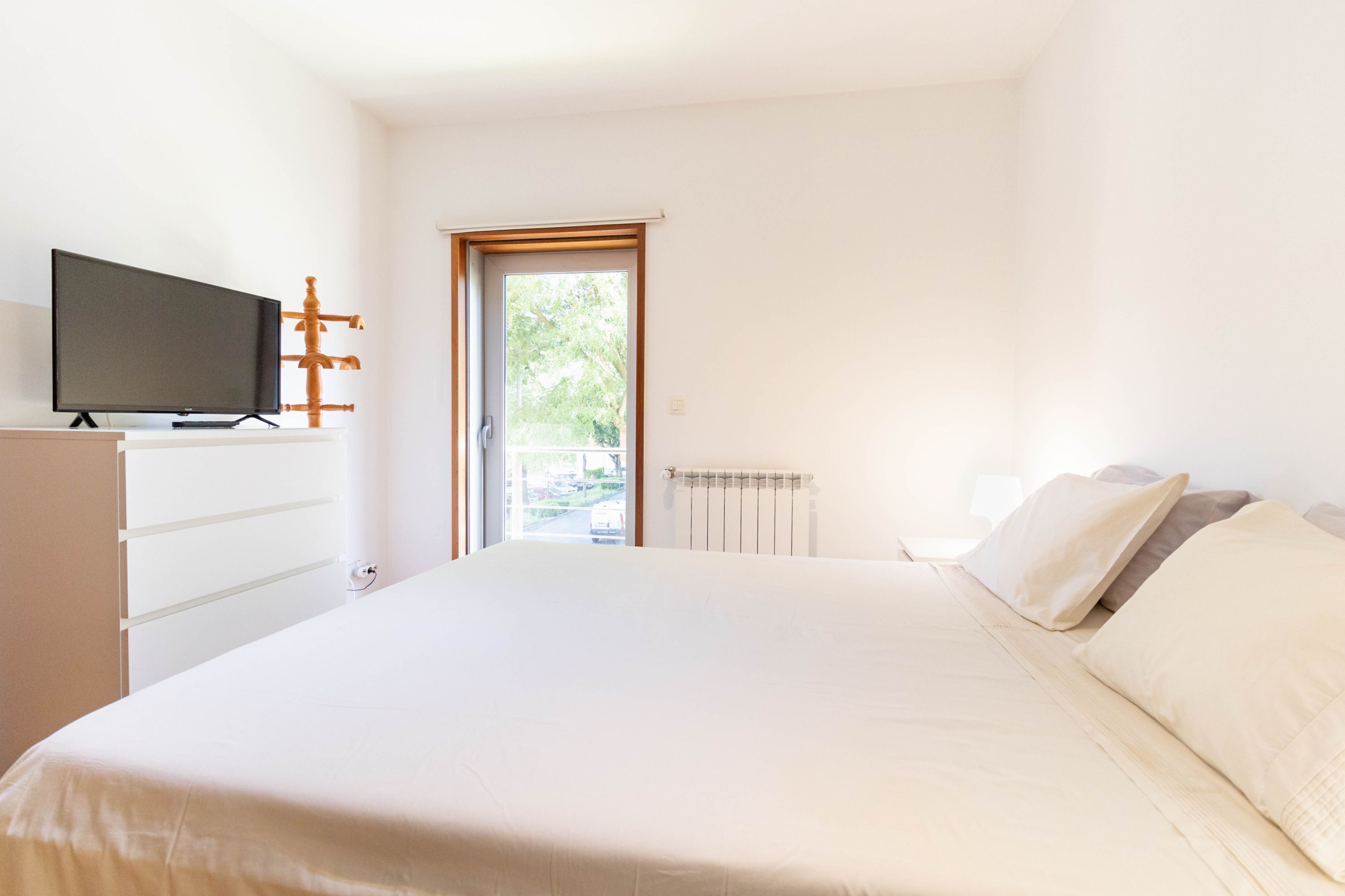 Une chambre à coucher avec un lit, un téléviseur posé sur une commode, un radiateur et une porte donnant sur un balcon.