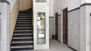Uma escada que dá acesso a um elevador, uma porta com painel de vidro e um corredor com paredes revestidas de azulejos e uma porta de madeira.