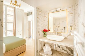 Une salle de bain moderne avec un grand miroir, un lavabo blanc, un pare-feu en marbre et un vase rempli de fleurs rouges.