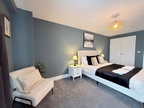 Une chambre à coucher comprenant un lit, des tables de chevet, une commode, une lampe, une œuvre d’art accrochée au mur et une porte.