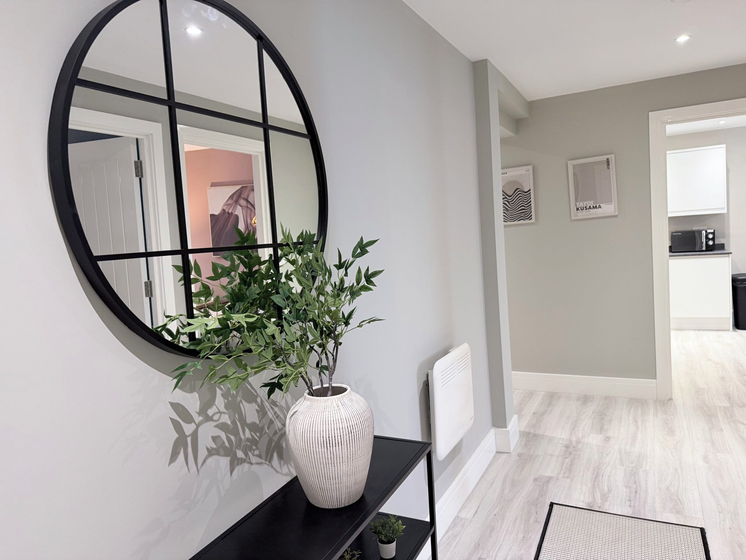 Et moderne interiør med et rundt spejl, en hvid vase med grønne planter, en sort hylde og et køkken i baggrunden.