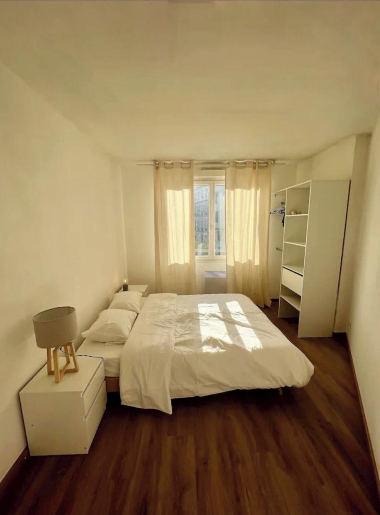 A bedroom with a bed, a bedside table, a lamp, a wardrobe, and a view of the outdoors through a window.