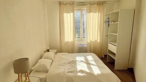 A bedroom with a bed, a bedside table, a lamp, a wardrobe, and a view of the outdoors through a window. - Apartment center, Timone/Stade vélodrome (Marseille)