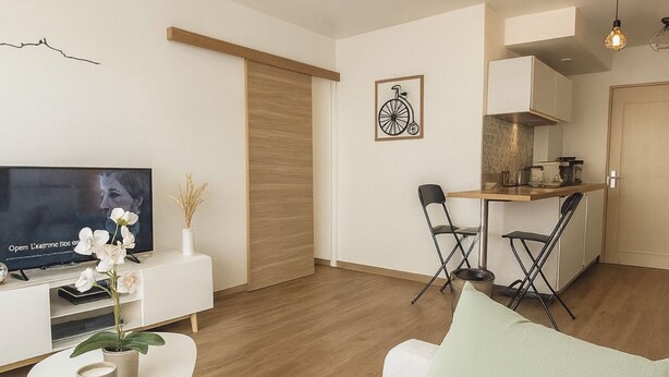 A modern living room with a sofa, a coffee table, and a TV. There is a dining area with chairs and a table. The room has wooden flooring and white walls.