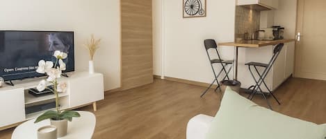 A modern living room with a sofa, a coffee table, and a TV. There is a dining area with chairs and a table. The room has wooden flooring and white walls.
