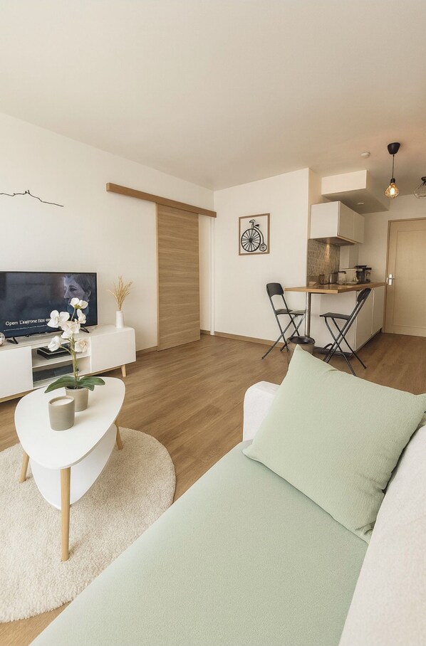 Una sala moderna con un sofá, una mesa de centro y un televisor. Hay una zona de comedor con sillas y una mesa. El piso es de madera y las paredes son blancas.