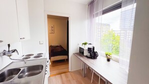 Cocina compacta con estufa, pileta y microondas. En la habitación contigua hay una cama y una ventana con vista a edificios y árboles.