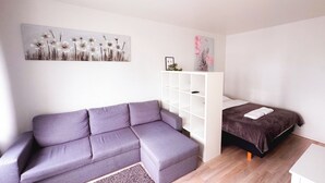 Un dormitorio moderno con un sofá seccional gris, un armario empotrado blanco, una cama individual con colcha marrón oscuro y dos cuadros colgados en la pared.