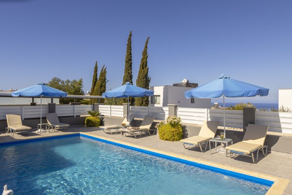 A rooftop pool area with lounge chairs and umbrellas, surrounded by cypress trees and a clear view of the ocean. - Lenias Sea View in Neo Chorio (Neo Chorio)