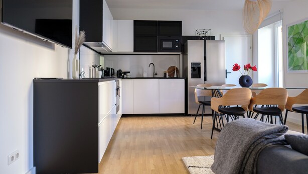 Cocina moderna con armarios blancos, refrigerador negro y comedor con sillas y mesa de madera.