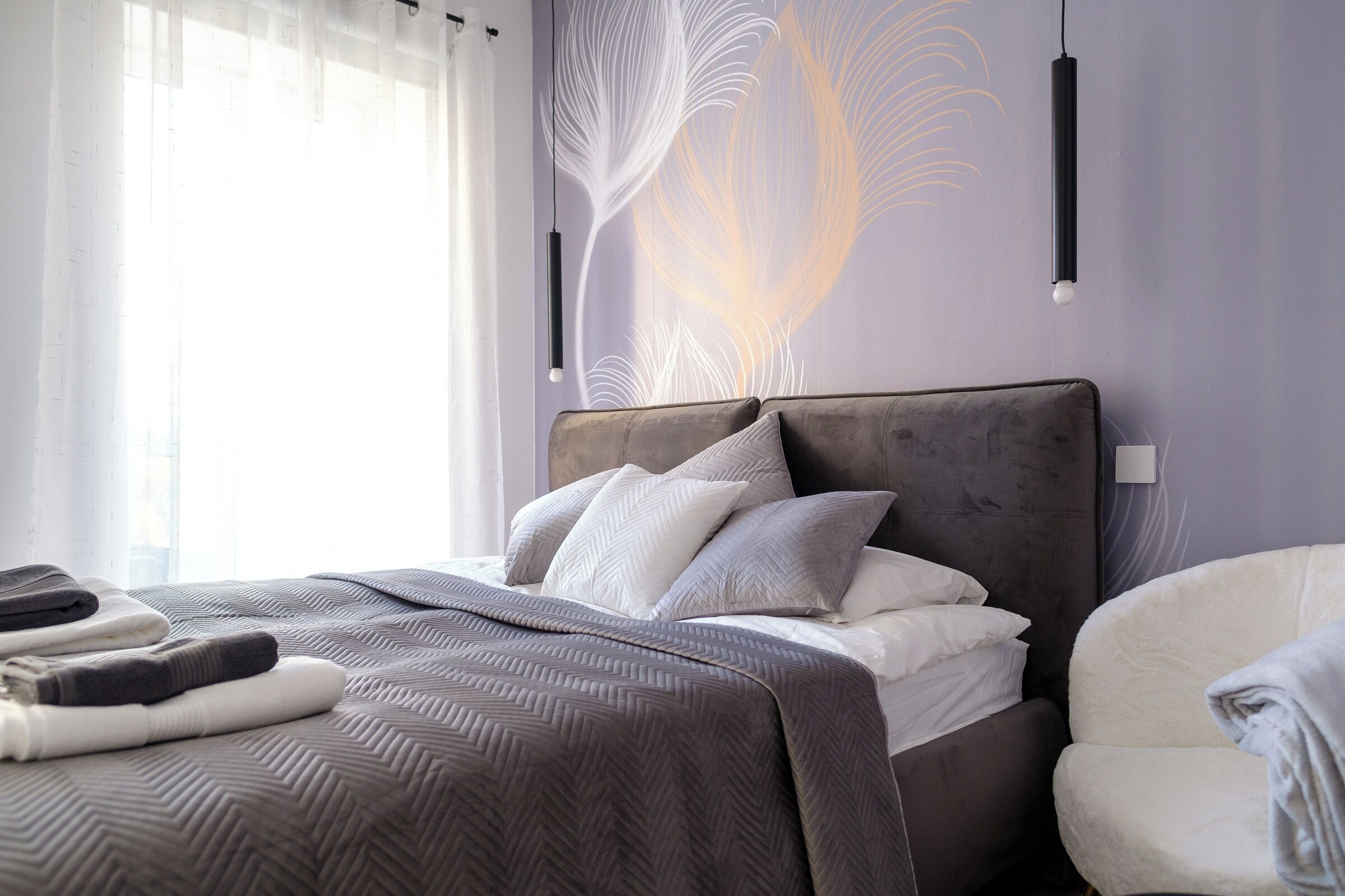 Ein ordentlich bezogenes Bett mit einer grauen Bettdecke im Fischgrätmuster und weißen Kissen, ein grau gepolsterter Sessel sowie eine Wand mit einem abstrakten, blattartigen Design.