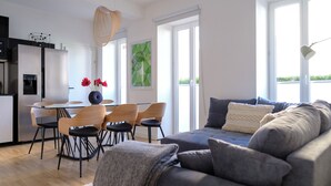 Una sala moderna con un sofá gris, una mesa de comedor con sillas y una zona de cocina con un refrigerador y dispensador de agua.