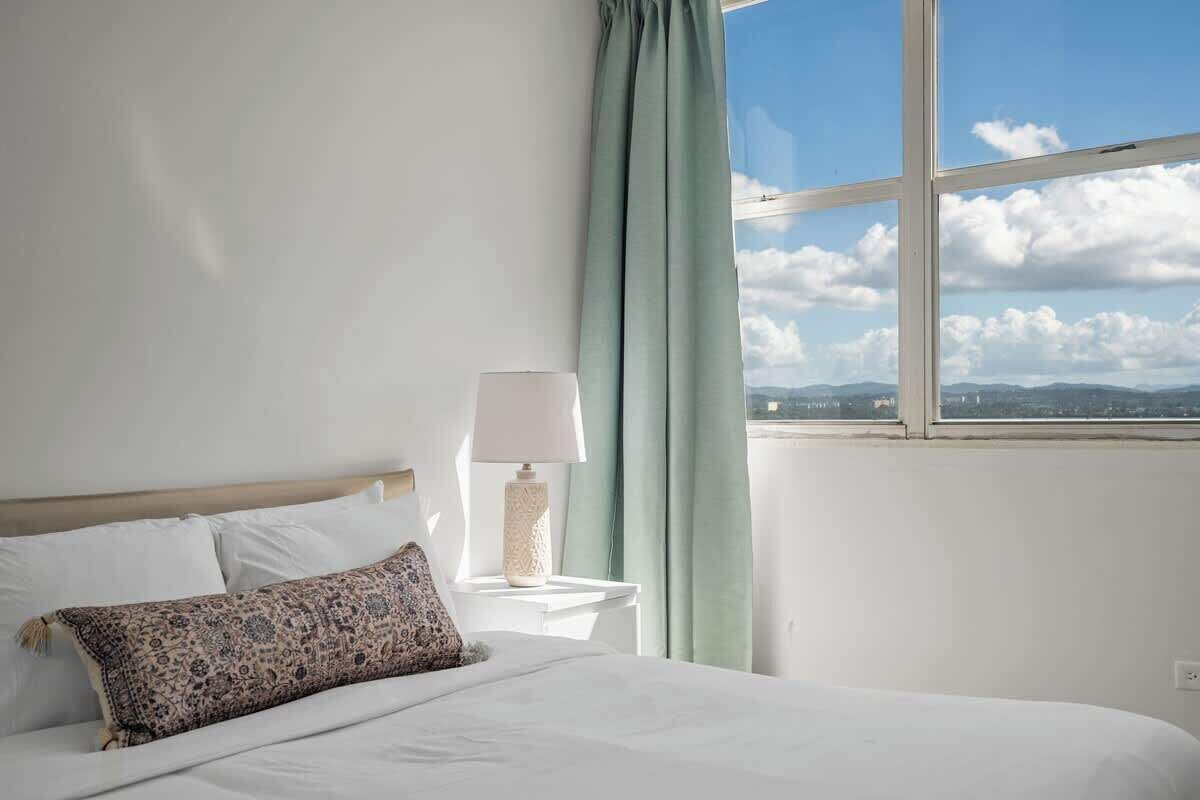 Ein Hotelzimmer mit Bett, Nachttisch mit Lampe und Blick auf Himmel und Berge durch ein Fenster.