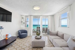 A spacious living room with a large window offering a view of the sea, a flat-screen TV mounted on the wall, a grey sofa, a black armchair, and a small table with a plant. - Beachfront | Pool | Ocean-View Balcony (Carolina)