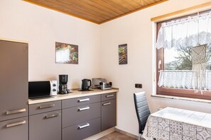 A kitchen with a microwave, coffee maker, toaster, and a table with a patterned tablecloth. There are two framed pictures on the wall, a window with lace curtains, and a black chair. - Holiday Apartment 'Göpfert' with Wi-Fi (Ebersburg-Weyhers)