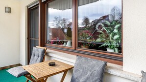 A balcony with a wooden table and chairs, a window with a view of a garden, and a potted plant on the windowsill.
