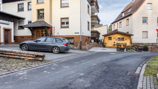 Una calle con un coche estacionado, una casa de madera y un edificio con balcón.