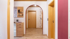 A hallway with wooden flooring, a wooden door at the end, a small cabinet with drawers, and a wall-mounted shelf with items.