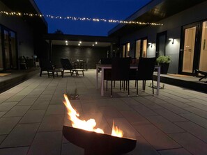 An outdoor patio area with a fire feature, seating, and string lights. - 5 Star Holiday Home in Vaeggerlose (Vaeggerlose)