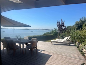 Een houten terras met een tafel en stoelen, met uitzicht op een rustig water waarin boten liggen.