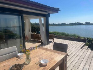 A wooden deck with a table set for two, overlooking a lake. - 5 Star Holiday Home in Martofte (Martofte)