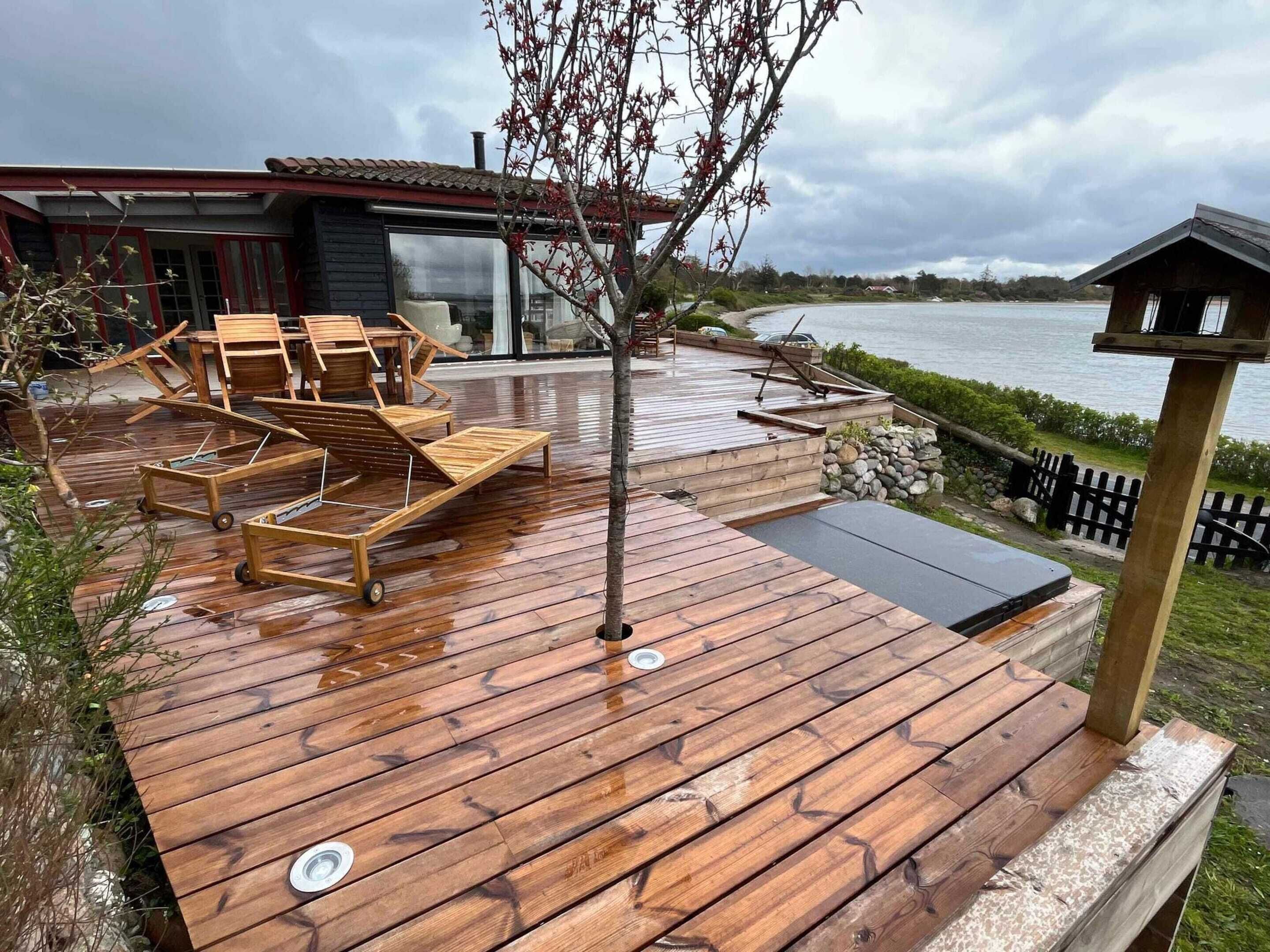 Une terrasse en bois avec des chaises longues et un petit arbre, donnant sur une étendue d’eau.