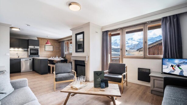 A modern living room with a fireplace, a dining area, and a kitchen. There is a large window with a mountain view. - Alaska 5 (Les Belleville)
