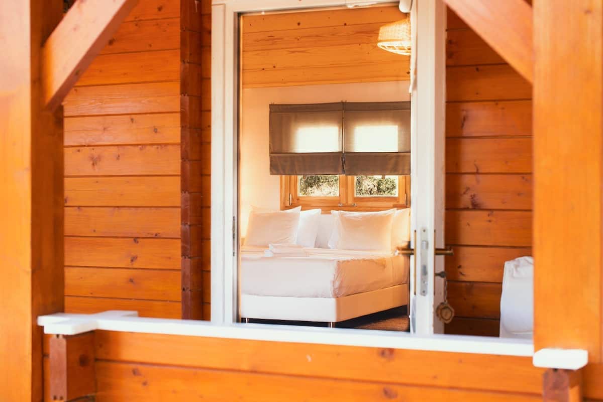 A wooden cabin with a bed in a loft-style room, a window with blinds, and a small shelf.