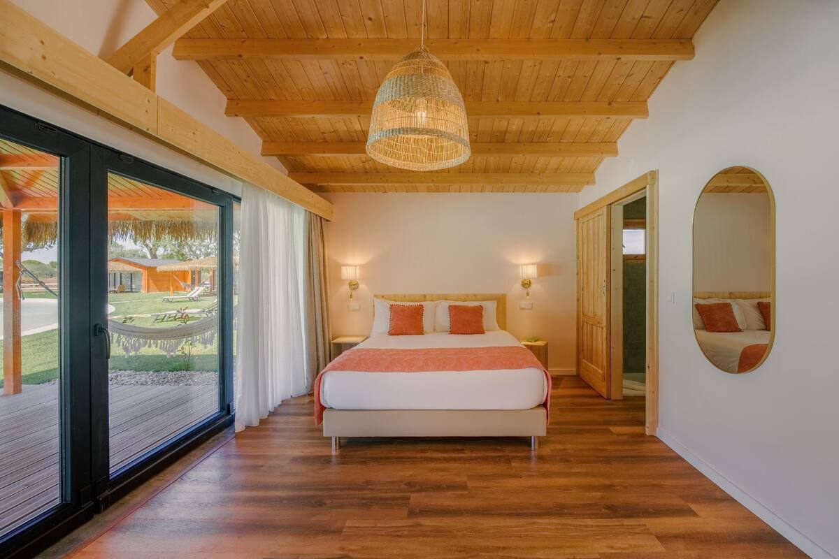 A bedroom with a large bed, wooden flooring, a hanging light fixture, and a view of an outdoor area.