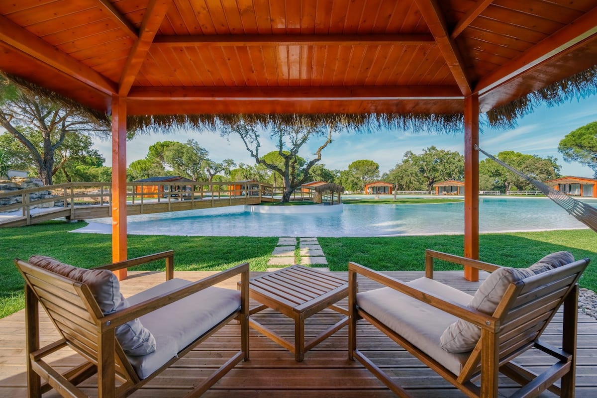 A wooden deck with two cushioned chairs and a small table, overlooking a swimming pool and a grassy area with trees.