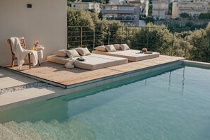 A poolside area with a wooden deck, two white sofas with cushions, a small table with a drink, and a chair with a towel.