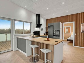 A modern kitchen with a large island, bar stools, a built-in oven, and a view of the outdoors. - 5 Star Holiday Home in Spottrup (Spøttrup)