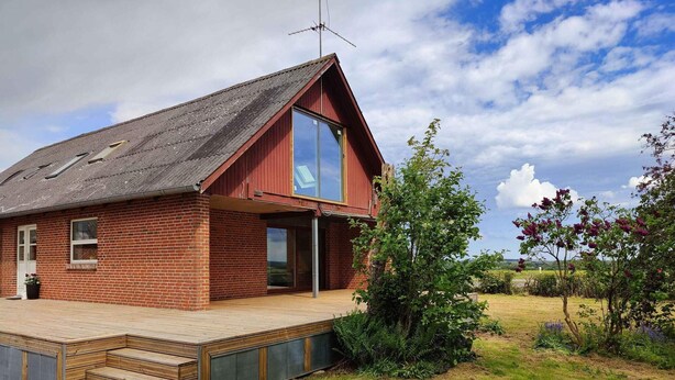 A red brick house with a large glass window, a wooden deck, and a garden with shrubs and flowers. - 5 Star Holiday Home in Galten (Galten)