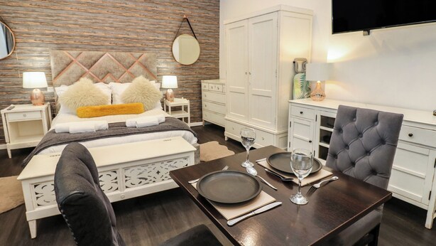 Une chambre moderne comprenant un lit, des tables de chevet, une télévision, un coin repas avec une table dressée pour deux personnes et une armoire.