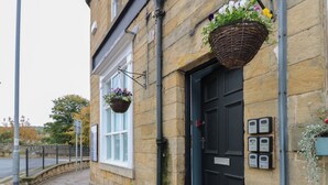 Un bâtiment en pierre avec une porte noire, une fenêtre ornée d’un panier suspendu rempli de fleurs et un petit jardinier avec des fleurs placé sur le trottoir.