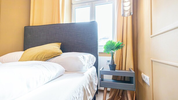 Un lit bien fait, avec du linge de lit blanc et un seul coussin jaune, une petite plante en pot sur une table de chevet et une fenêtre avec des rideaux.