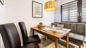 Ein Essbereich mit einem Holztisch, zwei schwarzen Stühlen, einer Weiden-Pendelleuchte, einem gerahmten Bild an der Wand und einem grauen Sofa mit Blick aus dem Fenster.